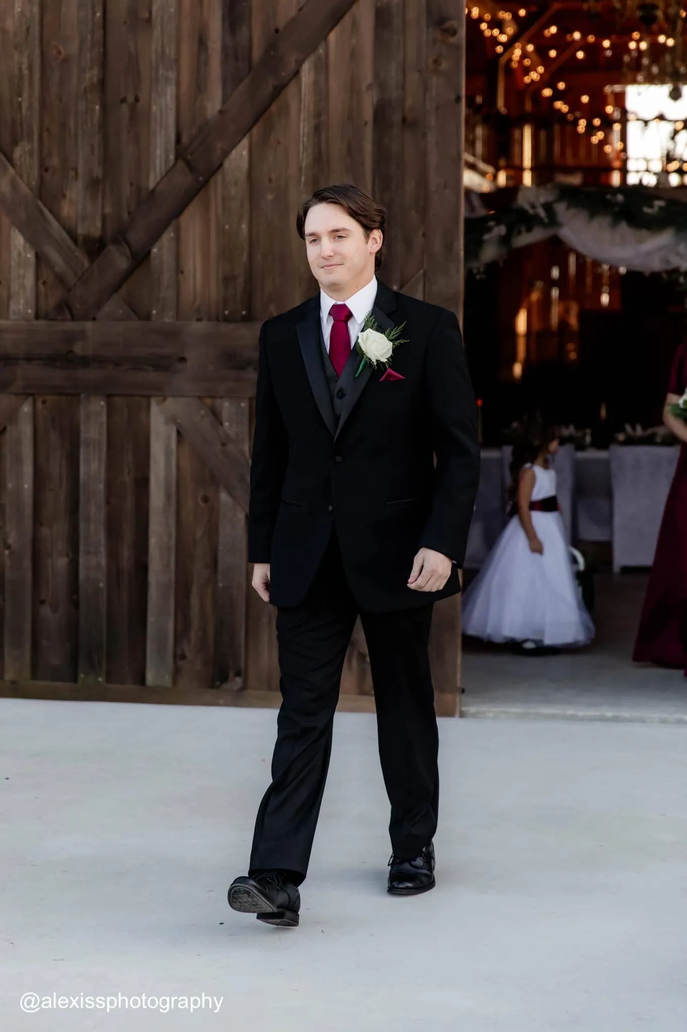 a man walking while wearing a black tuxedo rental with a red tie