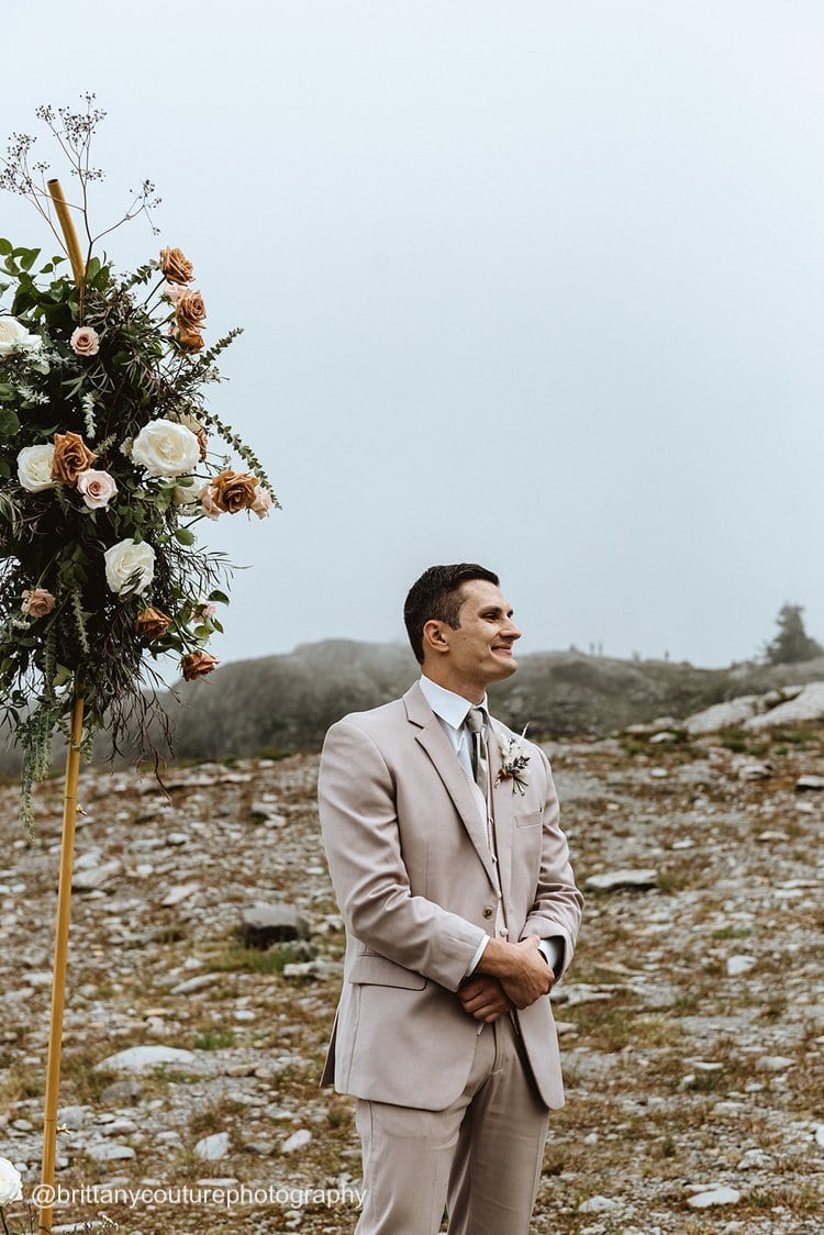 a groom wearing a tan suit rental waiting for his bride outside in a rocky mountain setting
