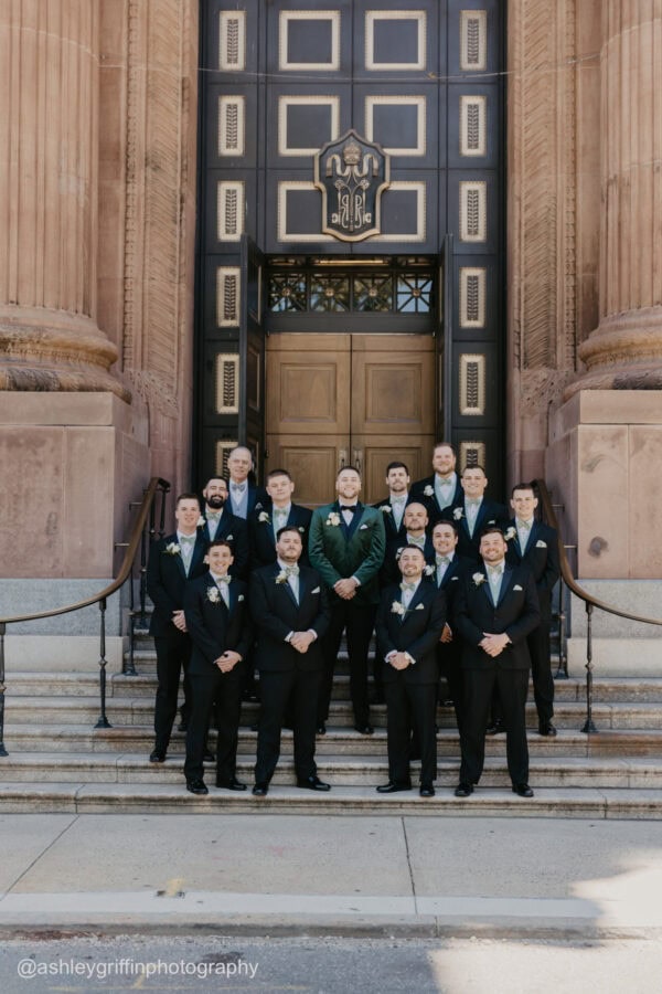 a groom in a green tuxedo standing on a stairway outside of a church surrounded by groomsmen