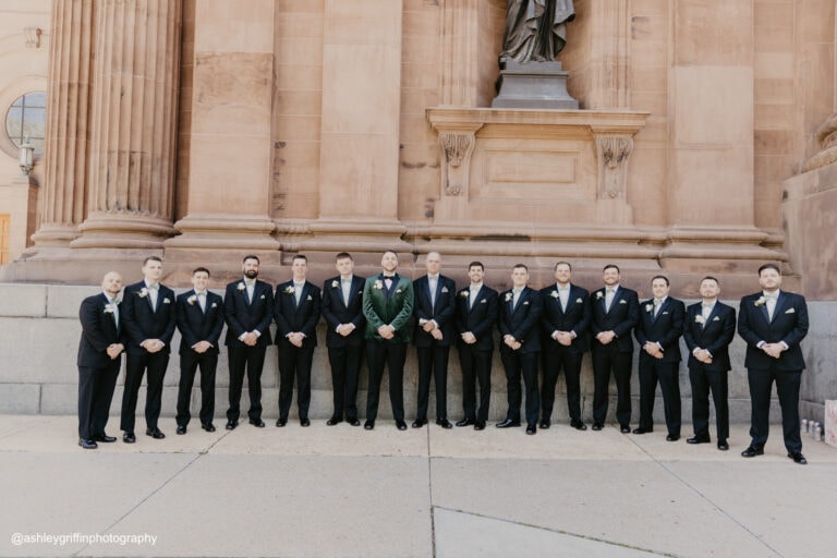 a groom wearing a green tuxedo rental standing outside of a church with his groomsmen