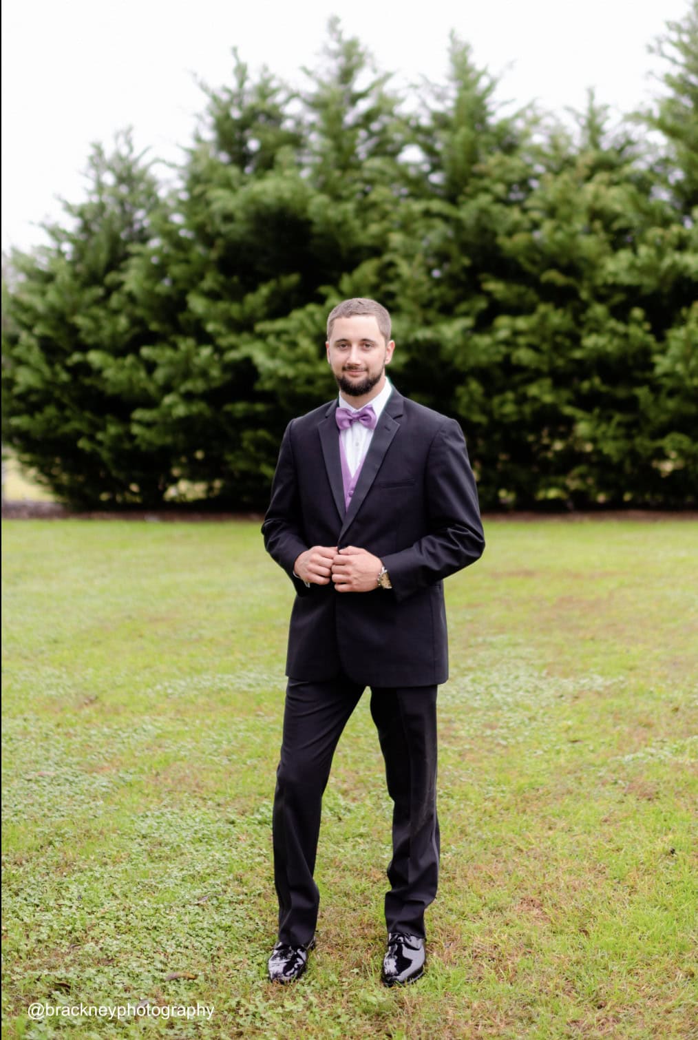 a groom standing outside wearing a black tuxedo rental with purple accessories
