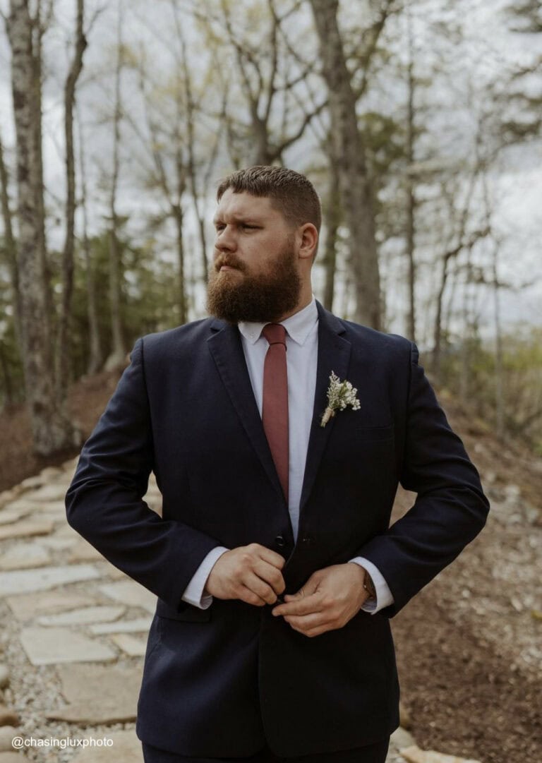 a groom wearing a navy suit rental by national tuxedo rentals buttoning his suit in a forest setting