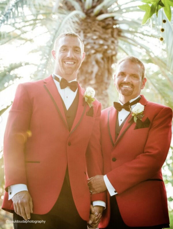 two men wearing red tuxedo rentals under a palm tree on wedding day