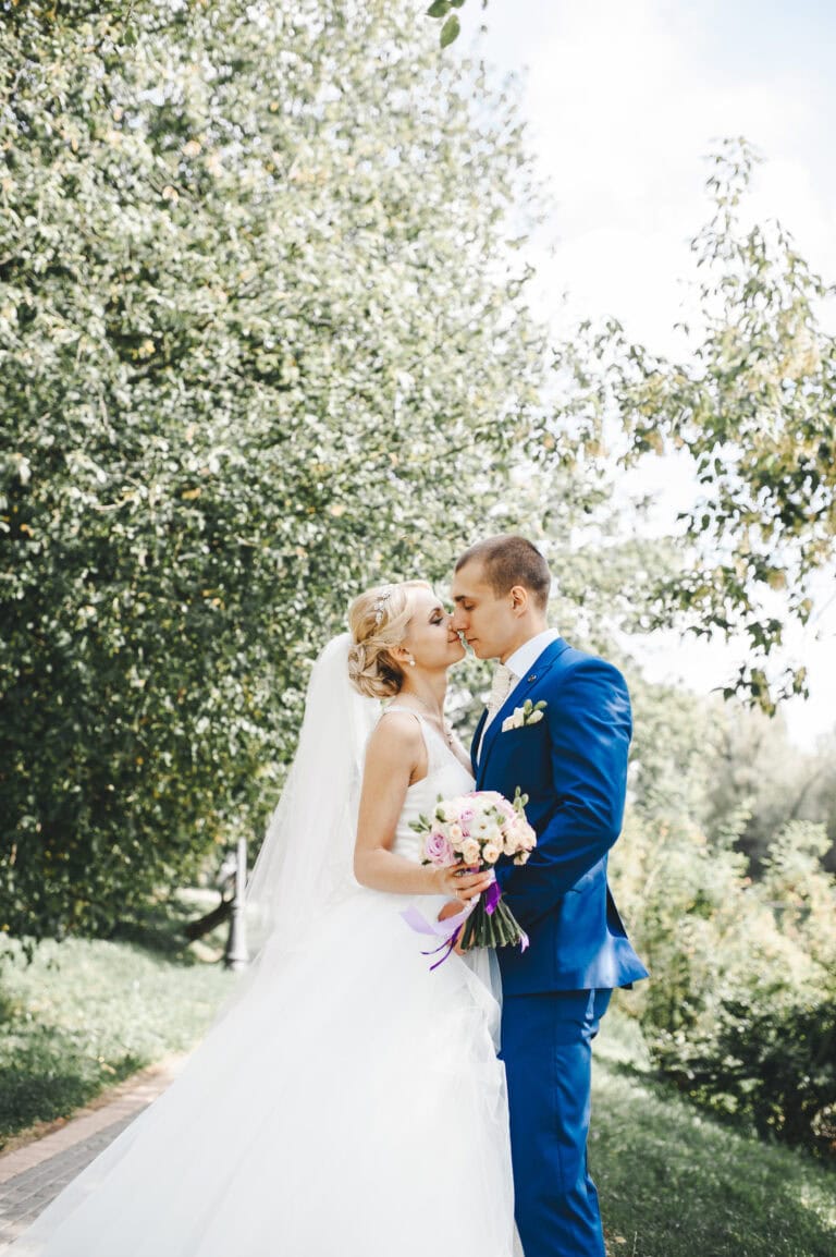 BeautifulWeddingCoupleInParkTheyKissAndHugEach | National Tuxedo Rentals man in blue suit rental with bride outdoors