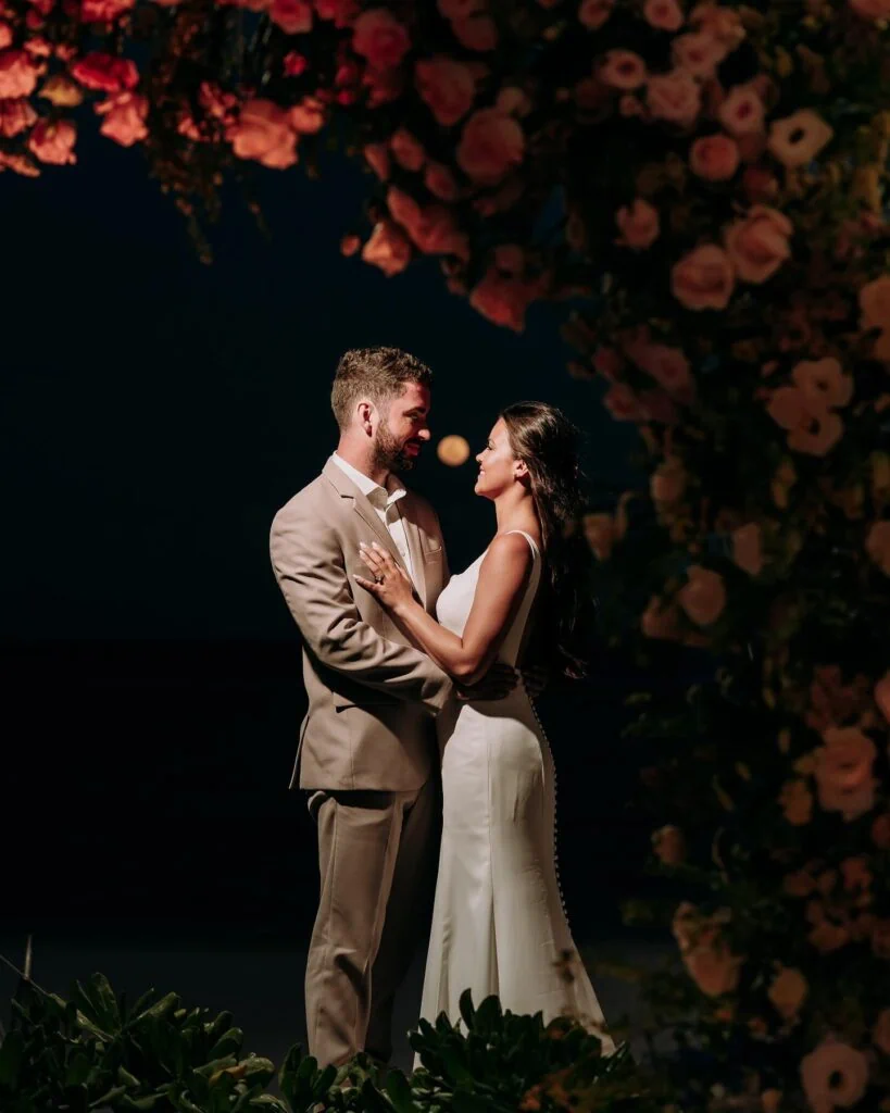 groom wearing tan suit with bride under flower arch in the dark