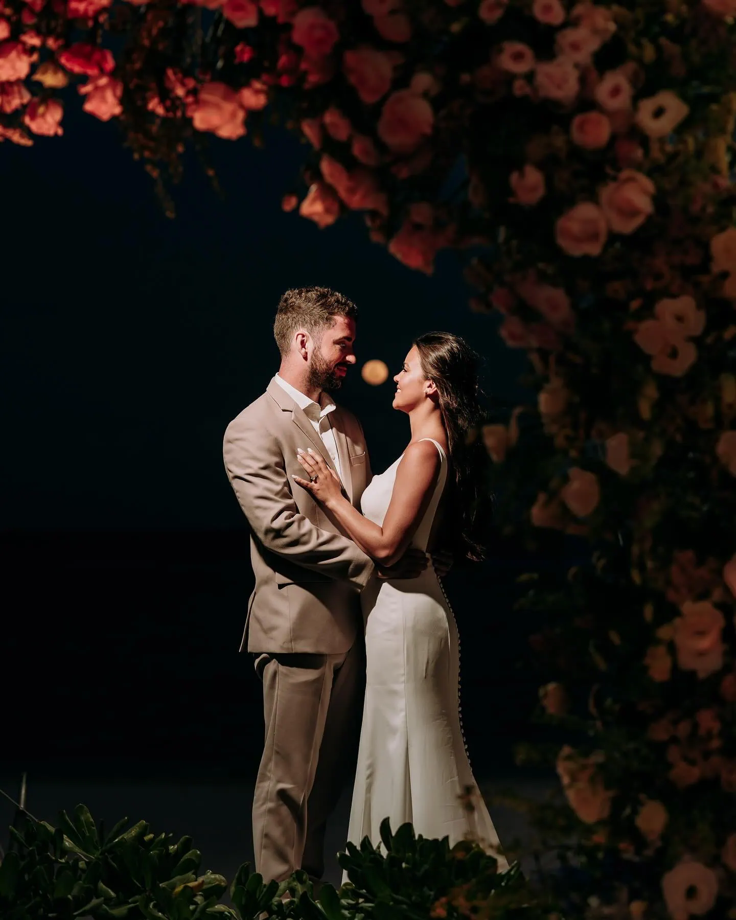 groom wearing tan suit with bride under flower arch in the dark