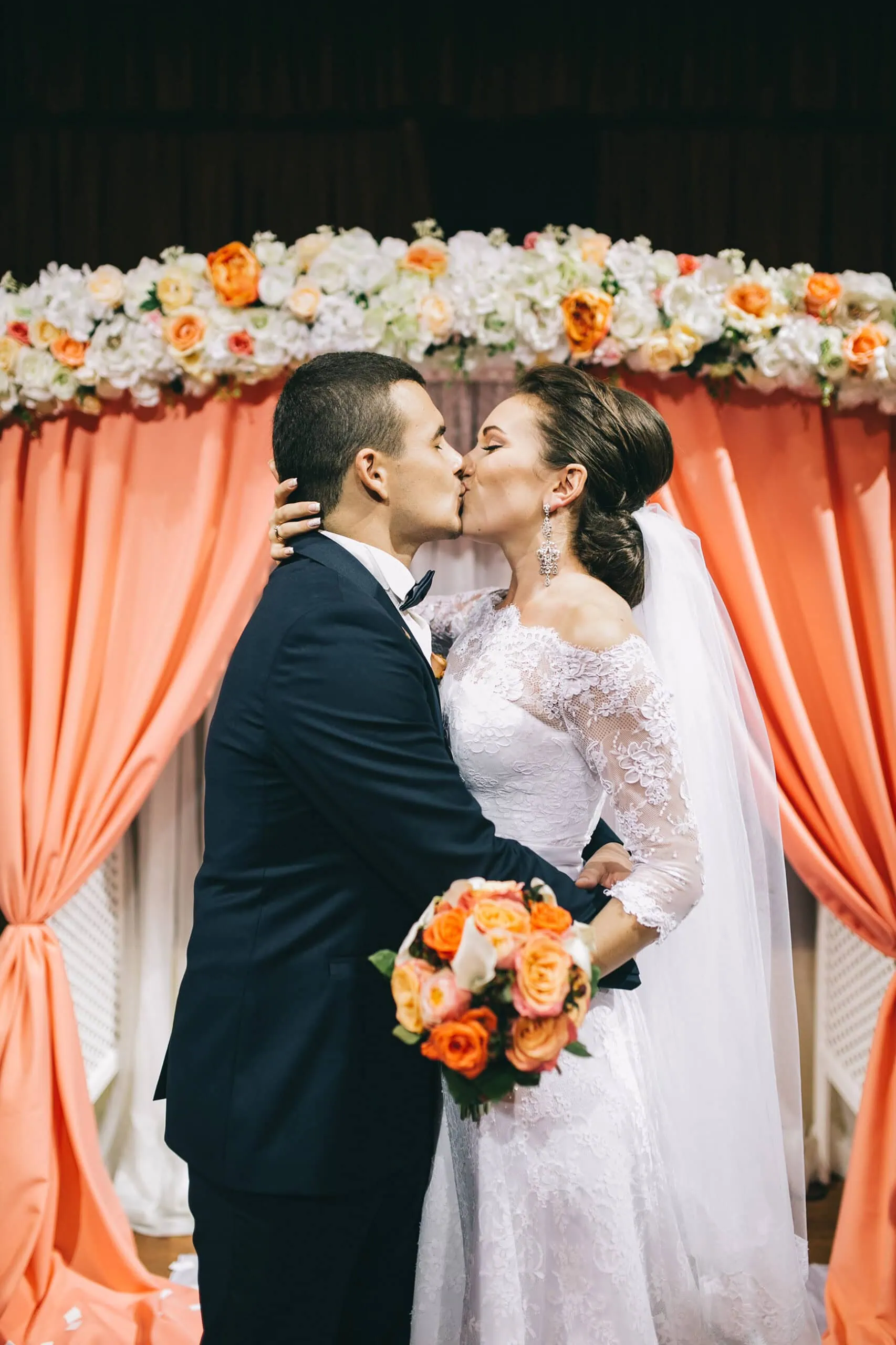 groom in navy suit kissing bride in front of orange flower arch