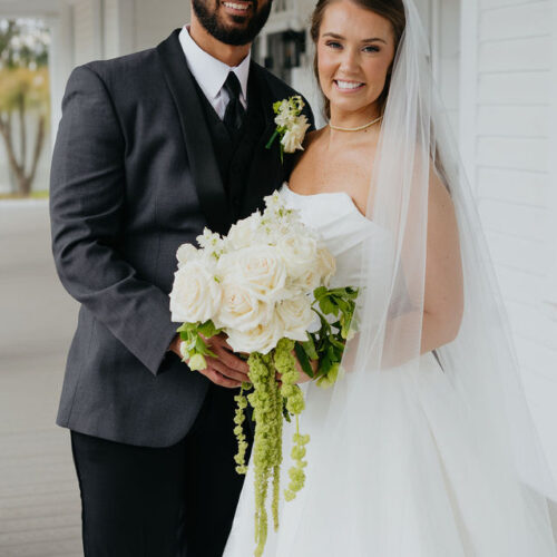 Bride in a white wedding dress and veil stands with a smiling groom in a dark suit on a porch, holding a bouquet of white roses.