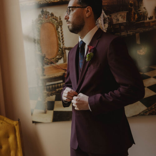 Man in a burgundy suit with boutonniere adjusts his jacket in a room filled with antique furniture and large paintings behind him, preparing for an event.