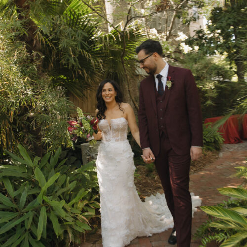 Bride in a white beaded gown and groom in a burgundy suit walk hand in hand along a brick garden path surrounded by tropical plants and white blossoms, smiling.