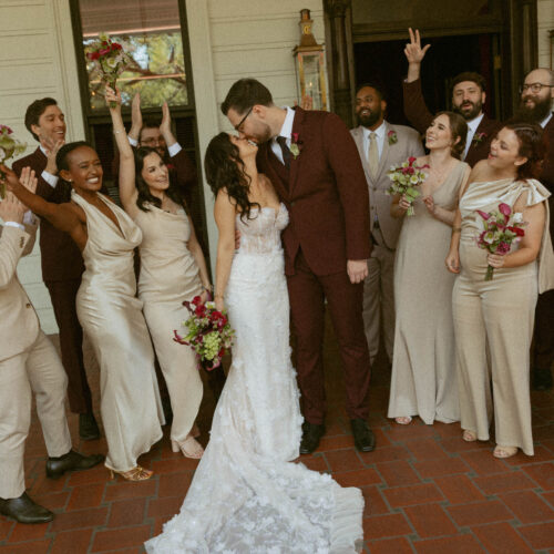 Bride and groom share a kiss as bridesmaids and groomsmen cheer around them on a brick porch.