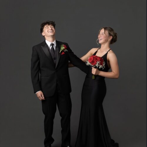 A smiling couple in formal attire posing together: man in a black tuxedo and tie, woman in a long black gown holding a bouquet of red roses.
