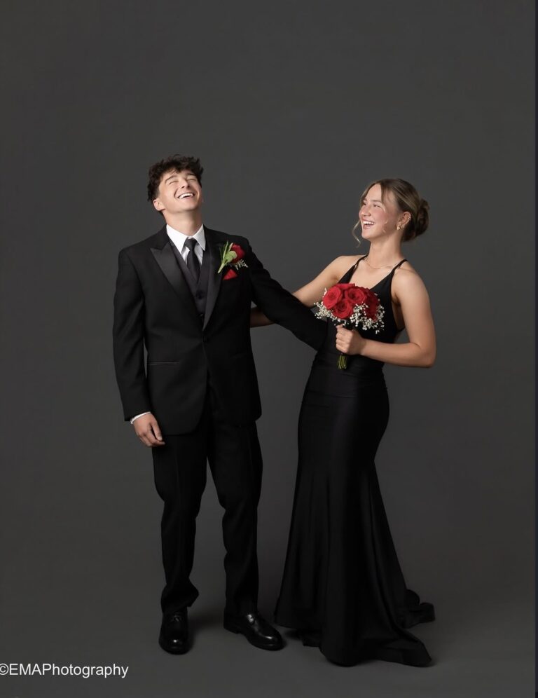 A smiling couple in formal attire posing together: man in a black tuxedo and tie, woman in a long black gown holding a bouquet of red roses.