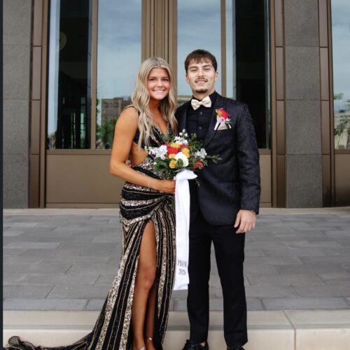 Couple in formal wear posing on steps outside a building; woman in a black and gold sequined gown holding a bouquet, man in a black suit with a cream bow tie.