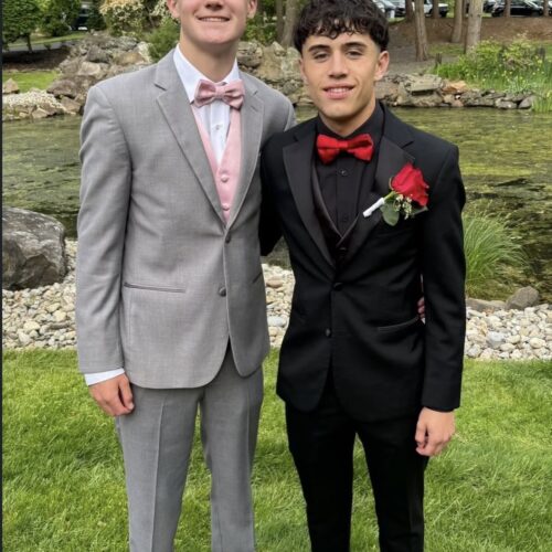 Two young men in formal suits posing outdoors by a pond and rocky garden setting, one in a gray suit with a pink bow tie and vest, the other in a black suit with a red bow tie and a red rose boutonniere.
