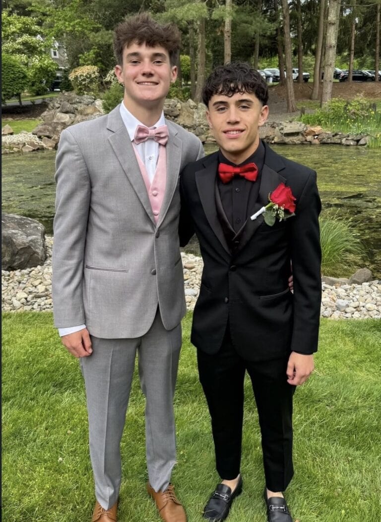 Two young men in formal suits posing outdoors by a pond and rocky garden setting, one in a gray suit with a pink bow tie and vest, the other in a black suit with a red bow tie and a red rose boutonniere.