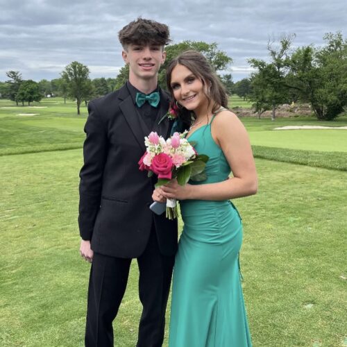 Couple dressed formally on a golf course: man in a black suit with teal bow tie and woman in a teal gown holding a pink bouquet.