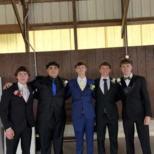 Five young men in suits standing side by side with arms around each other in a indoor, wooden-paneled space, one wearing a blue suit and brown shoes.