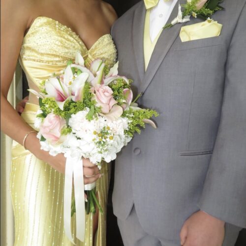 Bride in a pale yellow, beaded gown holds a pink-and-white bouquet; groom in a gray suit with yellow accents stands beside her.