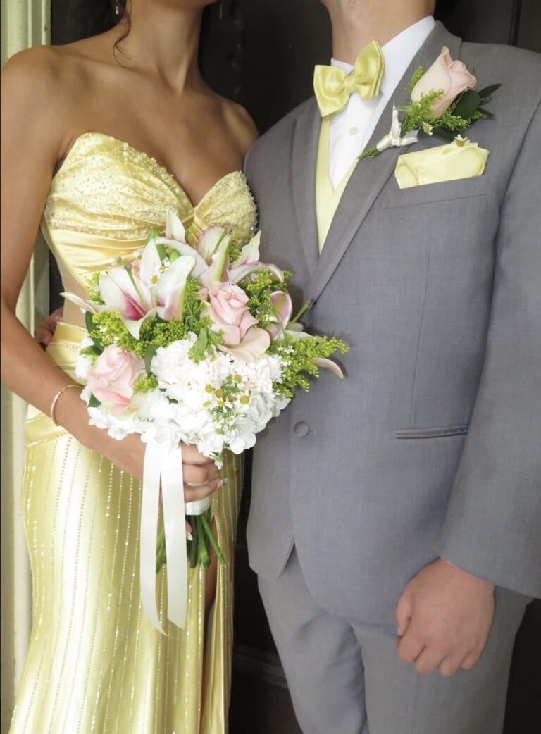 Bride in a pale yellow, beaded gown holds a pink-and-white bouquet; groom in a gray suit with yellow accents stands beside her.