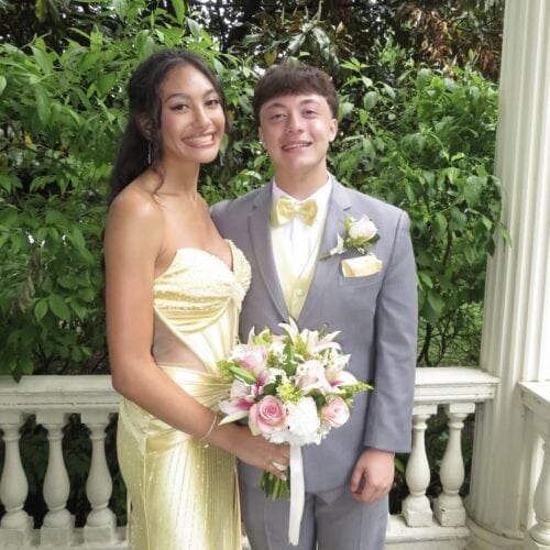 Smiling couple in formal attire outdoors; woman in pale yellow gown holding a pink bouquet, man in a gray suit with a yellow bow tie.