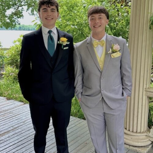 Two young men in formal suits posing on a wooden deck with greenery in the background; both wear boutonnieres and smiles.
