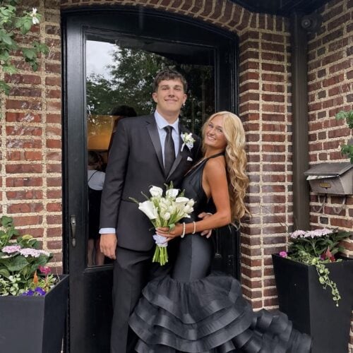 Couple in formal wear posing at a brick doorway: man in a black suit with boutonniere, woman in a black tiered gown holding a white bouquet.