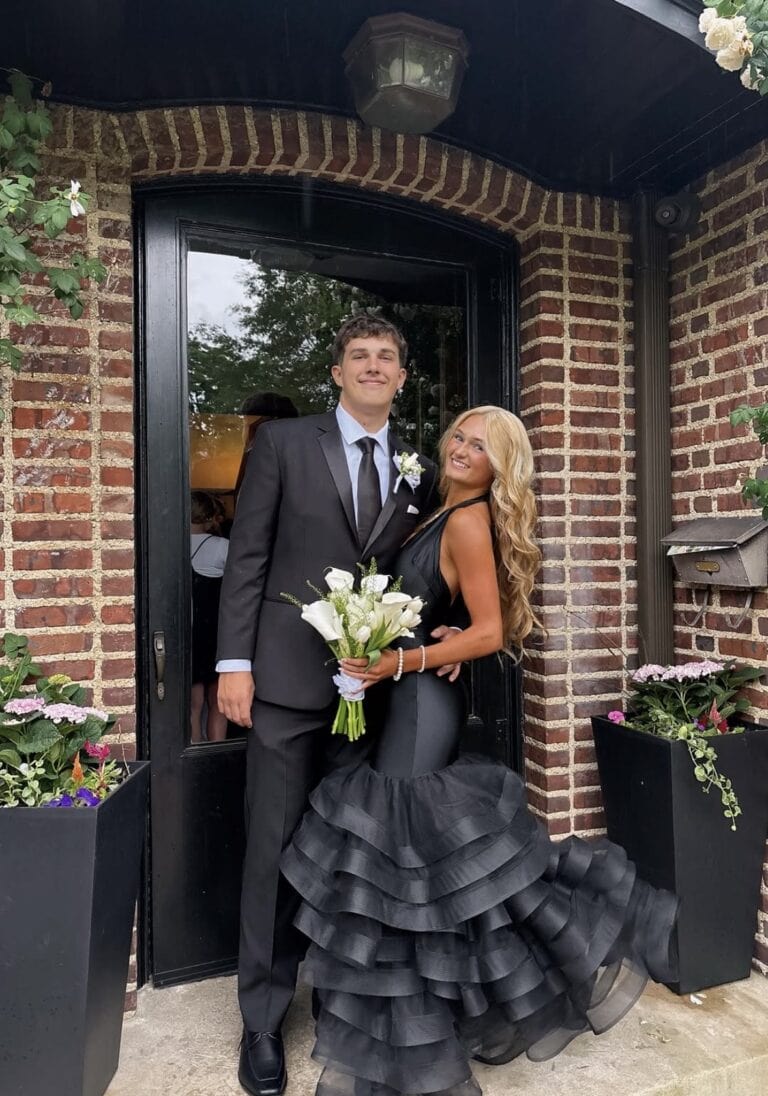 promcouple | National Tuxedo Rentals Couple in formal wear posing at a brick doorway: man in a black suit with boutonniere, woman in a black tiered gown holding a white bouquet.