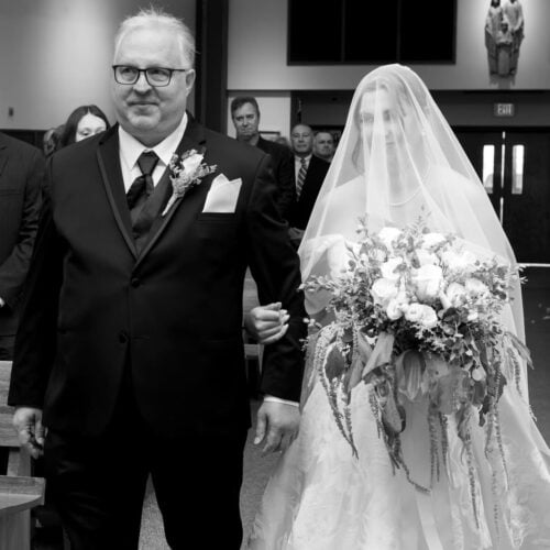 Man in a black tuxedo escorts a bride down the aisle; she wears a lace wedding gown with a veil and holds a large bouquet of flowers in front.