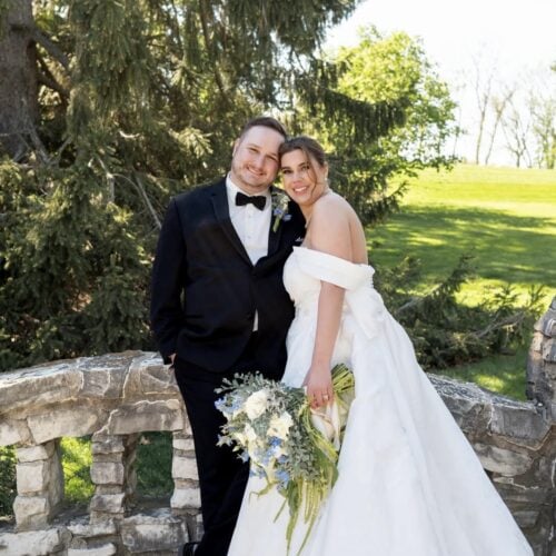 Bride in a white off-shoulder wedding gown holds a bouquet beside her groom in a black tuxedo on a stone railing outdoors.