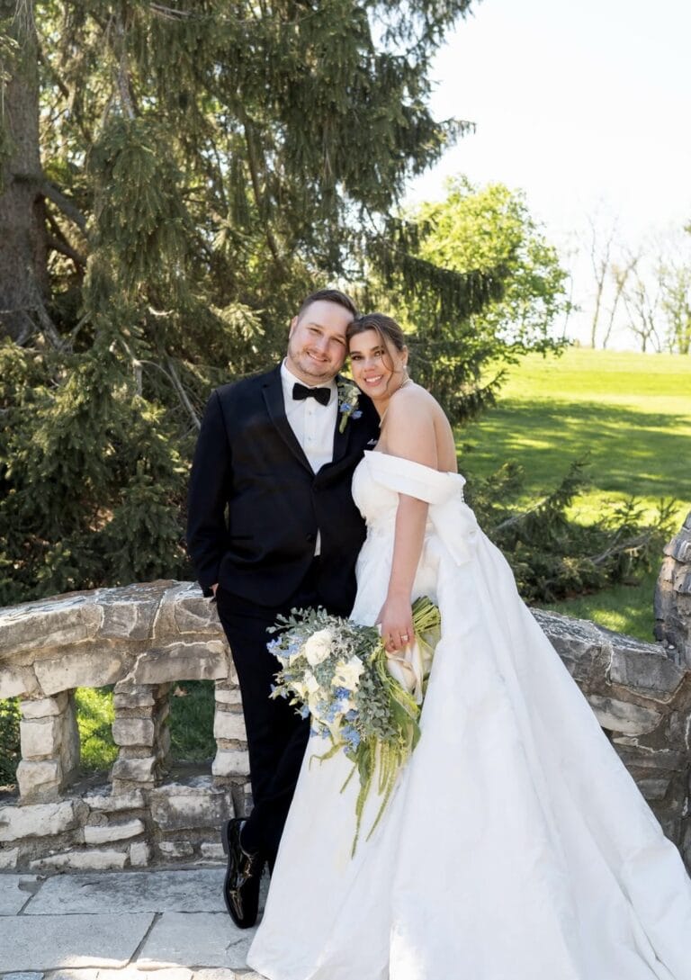Bride in a white off-shoulder wedding gown holds a bouquet beside her groom in a black tuxedo on a stone railing outdoors.