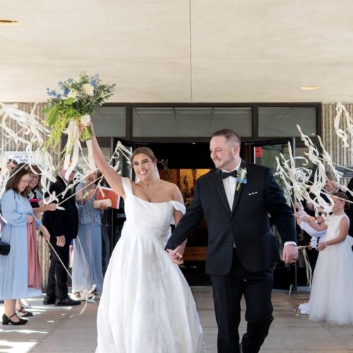 Bride in a white wedding gown raises her bouquet as she and the groom walk hand-in-hand through a streamered exit amid cheering guests outside a building.|Informative alt text for a wedding exit photo with couple and guests cheering at doorway, bride lifting bouquet, groom in tuxedo