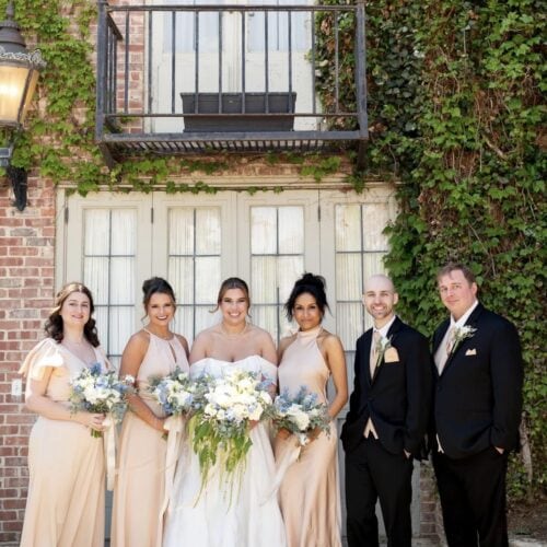 Wedding party posing outdoors: bride in white gown flanked by bridesmaids in peach dresses and two groomsmen in black suits with boutonnieres, ivy-covered brick building background