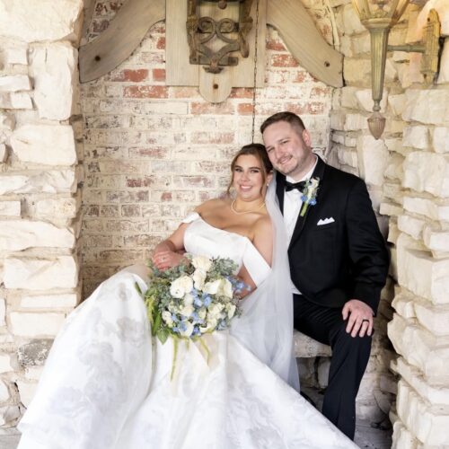 Bride and groom pose in a stone alcove, the bride in an off-shoulder white wedding gown holding a bouquet, the groom in a black tuxedo with a boutonniere.