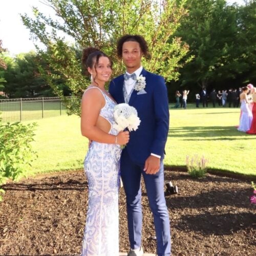 Couple in formal attire posing outdoors; woman in a light blue lace gown holding a white bouquet, man in a navy suit with a boutonniere against a sunny garden backdrop.