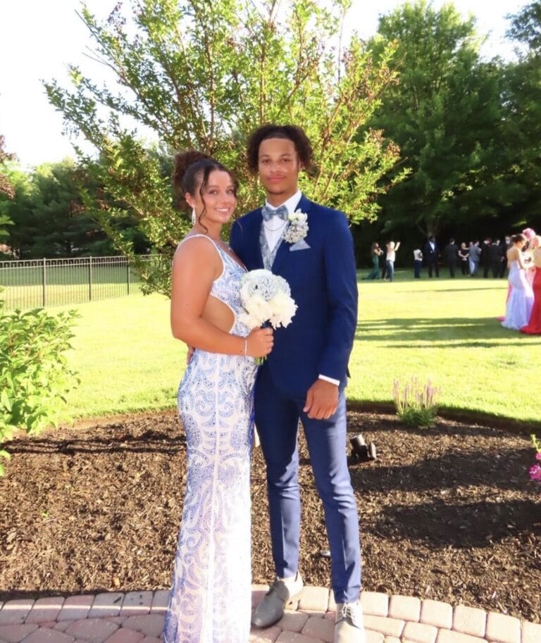 cobaltbluesuitprom | National Tuxedo Rentals Couple in formal attire posing outdoors; woman in a light blue lace gown holding a white bouquet, man in a navy suit with a boutonniere against a sunny garden backdrop.