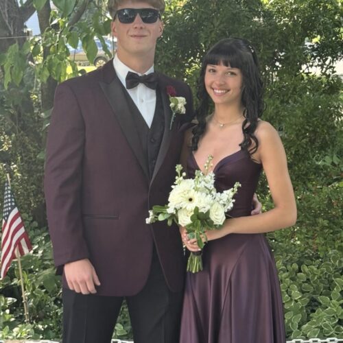 Couple in formal wear outdoors: man in a burgundy suit with bow tie and sunglasses, woman in a burgundy gown holding a white bouquet.