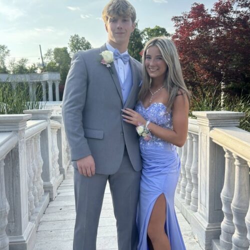 Young couple in formal wear posing on a marble railing outdoors; man in gray suit with a light blue vest and bow tie, woman in a lavender gown with a slit.