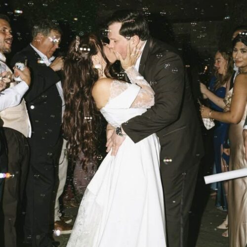 Bride and groom share a kiss while guests celebrate around them at a wedding reception with bubbles in the air.