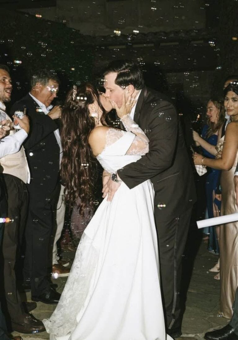 Bride and groom share a kiss while guests celebrate around them at a wedding reception with bubbles in the air.