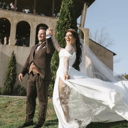 Bride in a white wedding dress and groom in a brown suit dancing outdoors as the veil flows behind them, under a clear blue sky.
