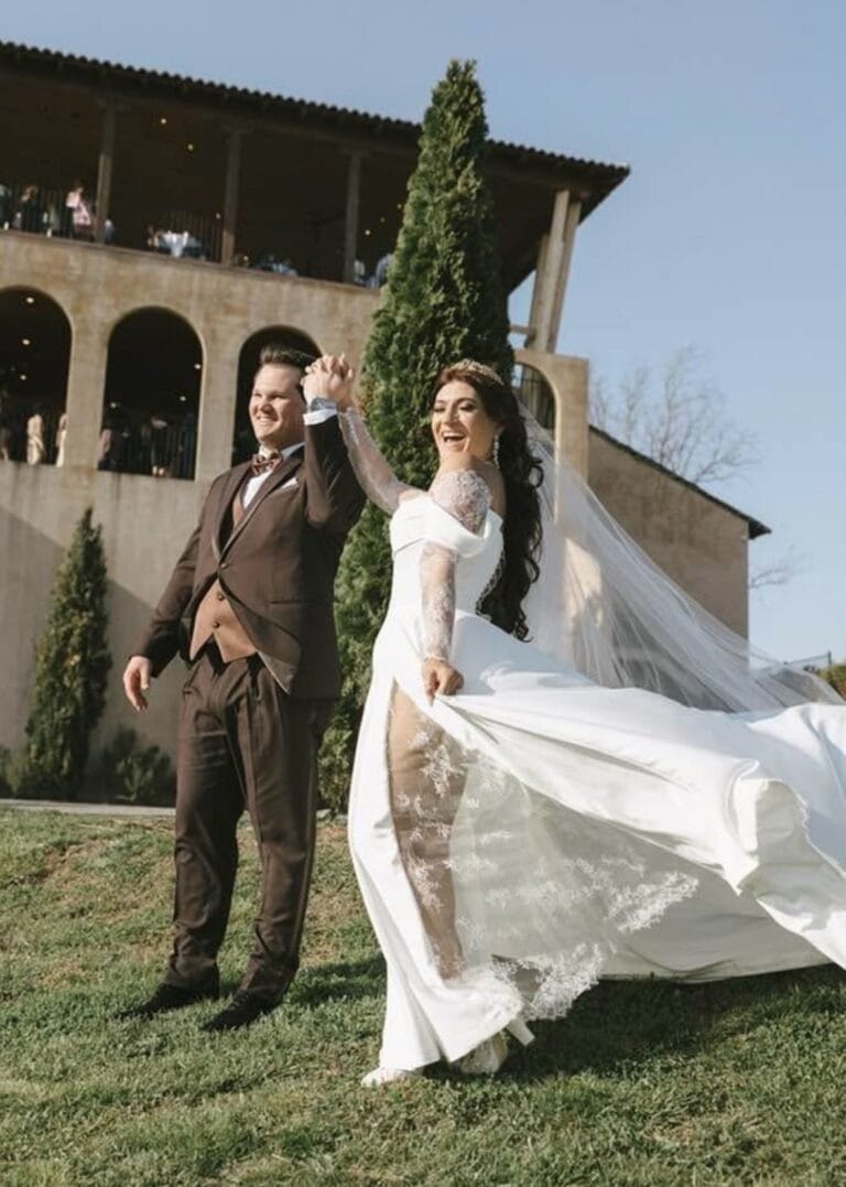 Bride in a white wedding dress and groom in a brown suit dancing outdoors as the veil flows behind them, under a clear blue sky.