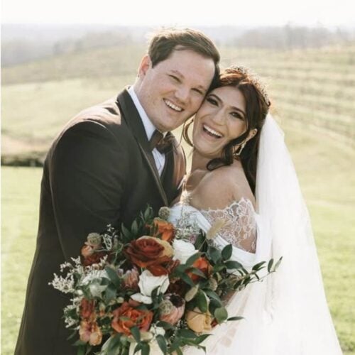 Bride and groom in wedding attire smile and hug, holding a large bouquet of orange, white flowers outdoors on a sunny day.