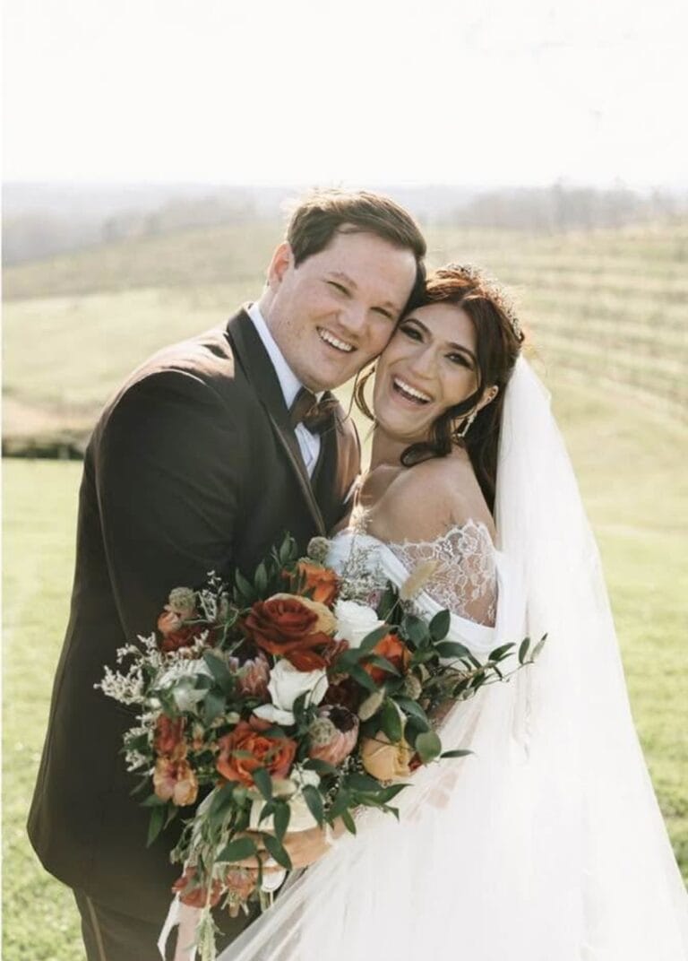 Bride and groom in wedding attire smile and hug, holding a large bouquet of orange, white flowers outdoors on a sunny day.
