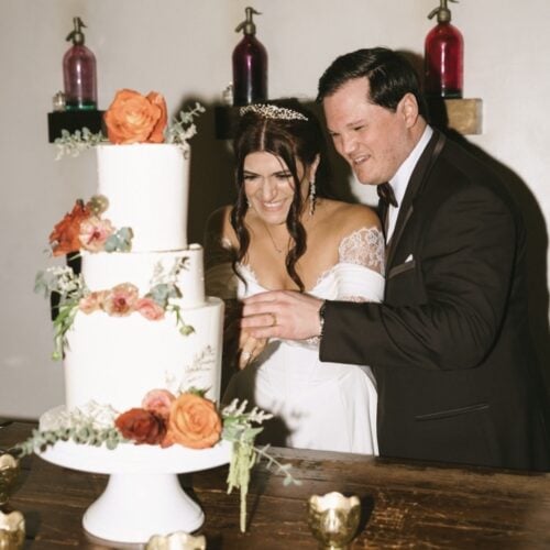 Bride and groom cutting a white tiered wedding cake adorned with orange and pink flowers, smiling at the ceremony.