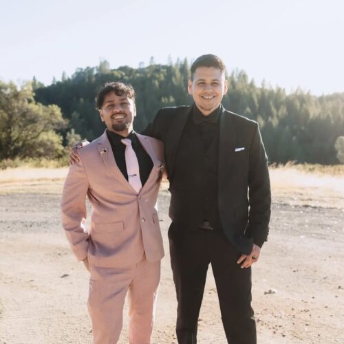 Two men in formal suits standing outdoors on a sunny day, smiling; one wearing a pink suit and pink tie, the other a black suit with a white pocket square.