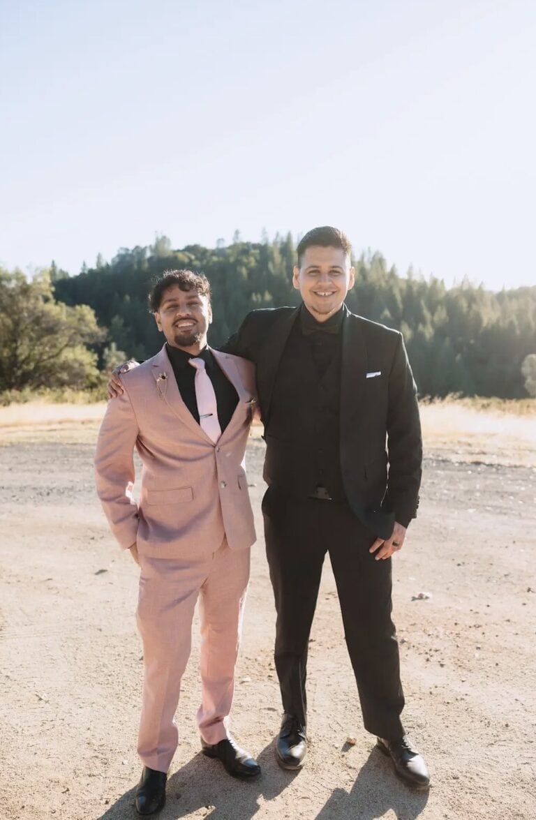 Two men in formal suits standing outdoors on a sunny day, smiling; one wearing a pink suit and pink tie, the other a black suit with a white pocket square.