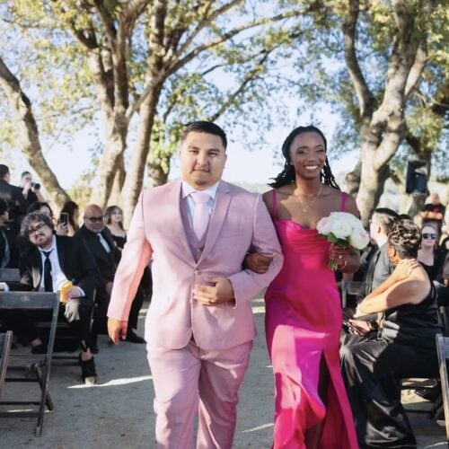 Man in a light pink suit walks arm-in-arm with a woman in a bright pink dress at an outdoor wedding aisle, holding a bouquet of white flowers.