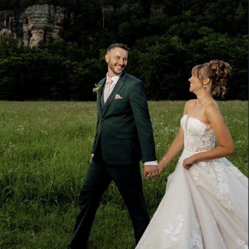 Bride in a white lace gown and groom in a dark green suit walk hand in hand across a grassy field, with trees behind them.