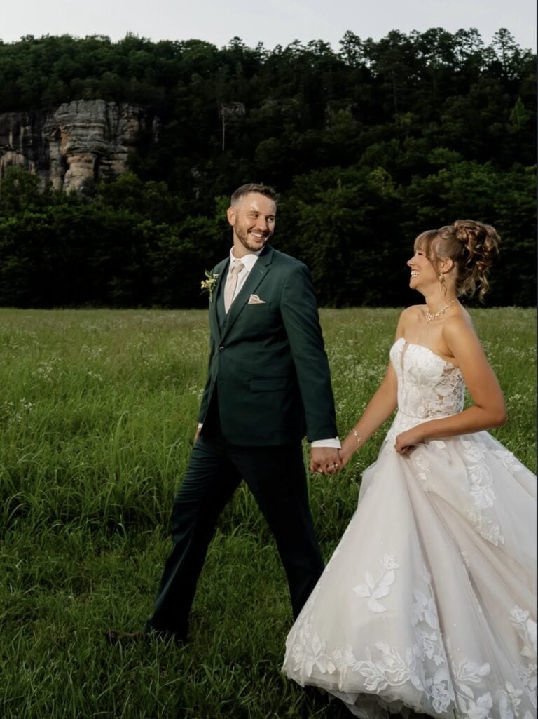 greensuit | National Tuxedo Rentals Bride in a white lace gown and groom in a dark green suit walk hand in hand across a grassy field, with trees behind them.